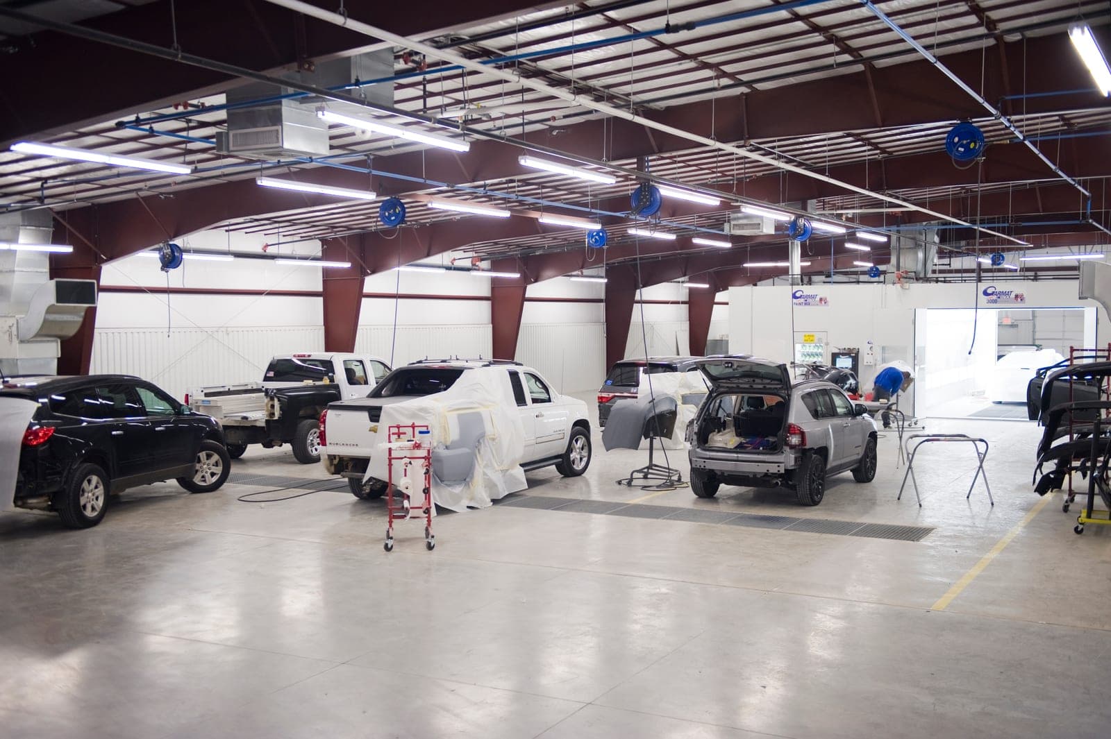 An open collision repair floor with vehicles at different stages of work — panels removed, one truck masked for paint — and the paint booth at the far end.